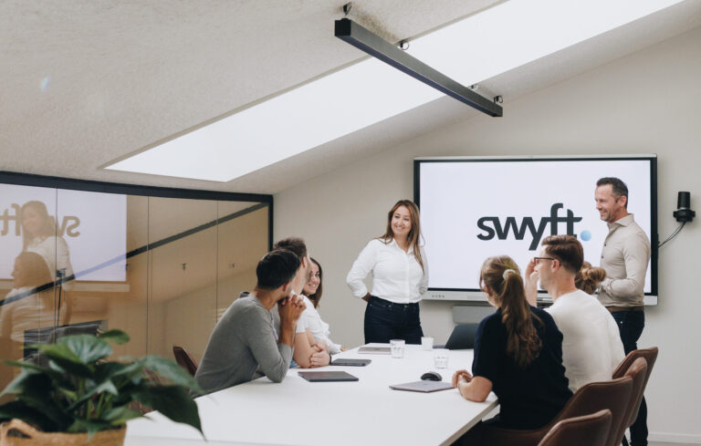 Vergaderruimte waarin een team rond een tafel zit terwijl twee collega’s een presentatie geven bij een scherm.